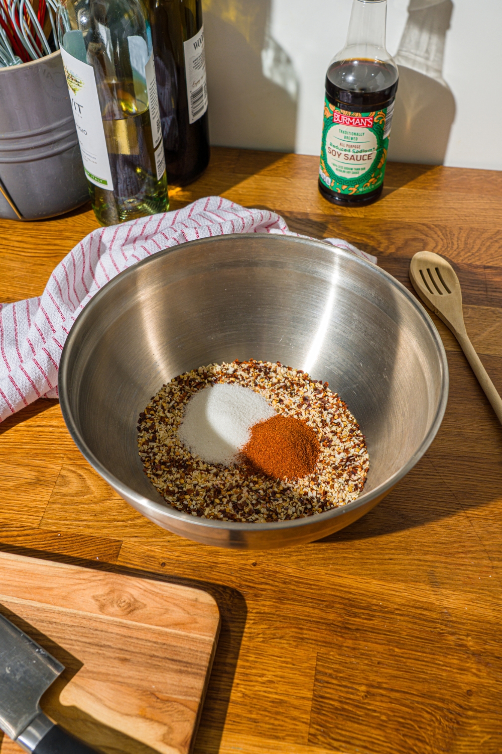 A large bowl with toasted sesame seeds, chili flakes, minced onion, and seasonings. The bowl is on a wooden counter with a wooden spoon and white striped napkin.
