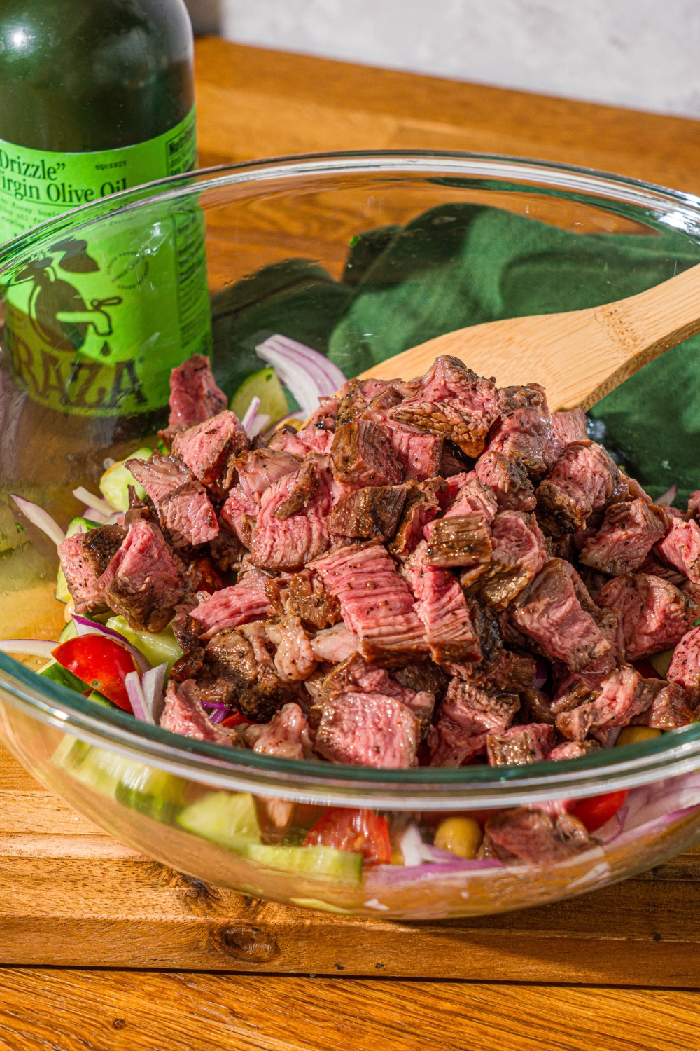 A glass bowl with sliced cucumbers, tomatoes, red onion, and chick peas with sliced grilled steak on top. A wooden spoon is stirring the mixture.