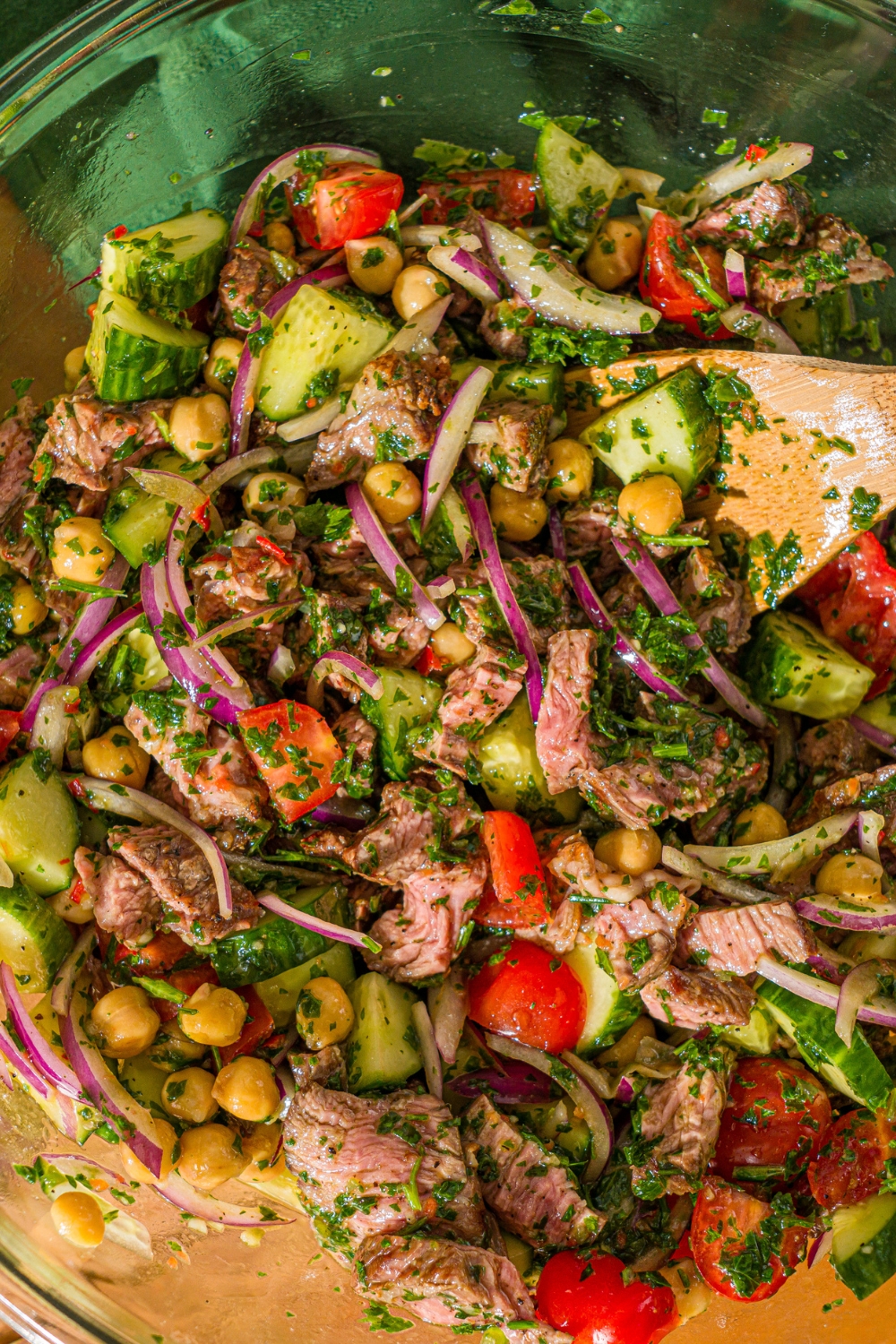 A glass bowl with steak, sliced cucumbers, tomatoes, red onion, and chick peas with chimichurri sauce. A wooden spoon is stirring the mixture.