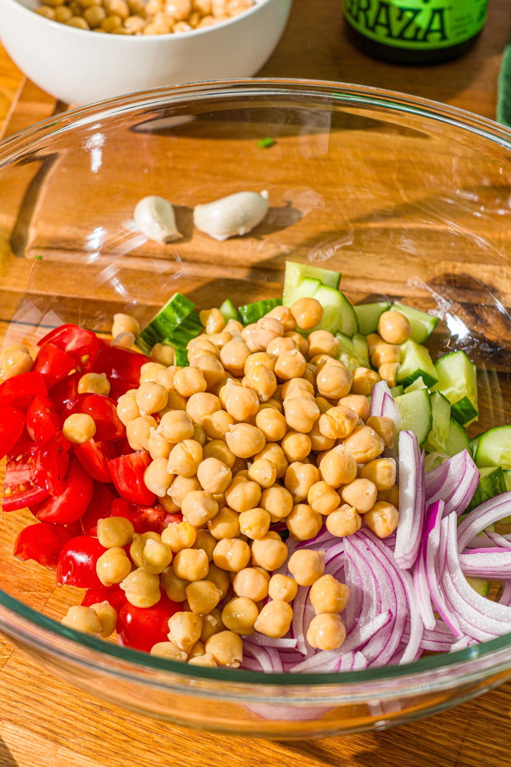 A glass bowl with sliced cucumbers, tomatoes, red onion, and chick peas. The bowl is on a wooden counter.
