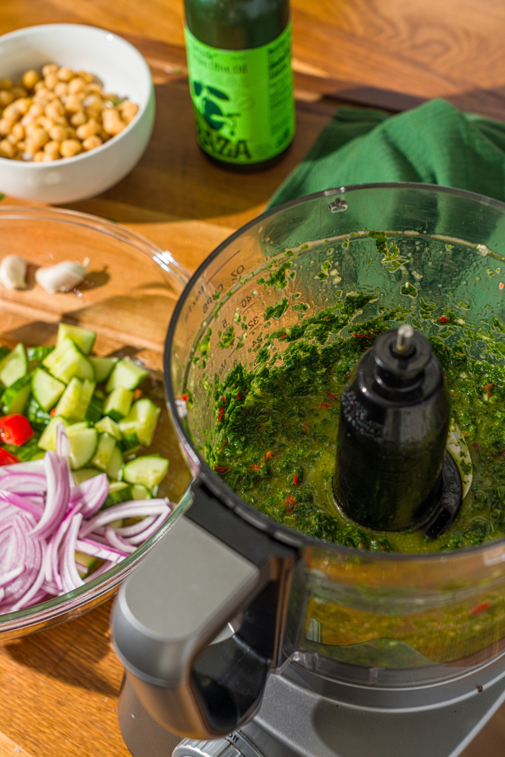 A food processor with chimichurri dressing. The food processor is on a wooden counter with a bowl of sliced vegetables.