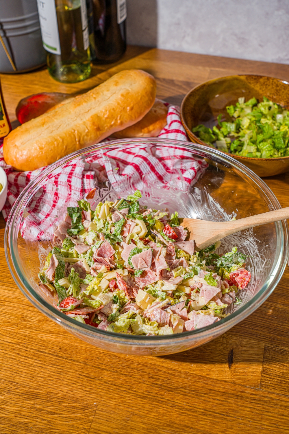 A glass bowl with a wooden spoon tossing ingredients to make chopped Italian sandwich including chopped deli ham, lettuce, salami, provolone, and vegetables in a dressing. The bowl is on a wooden counter with a red striped napkin and a hoagie roll.