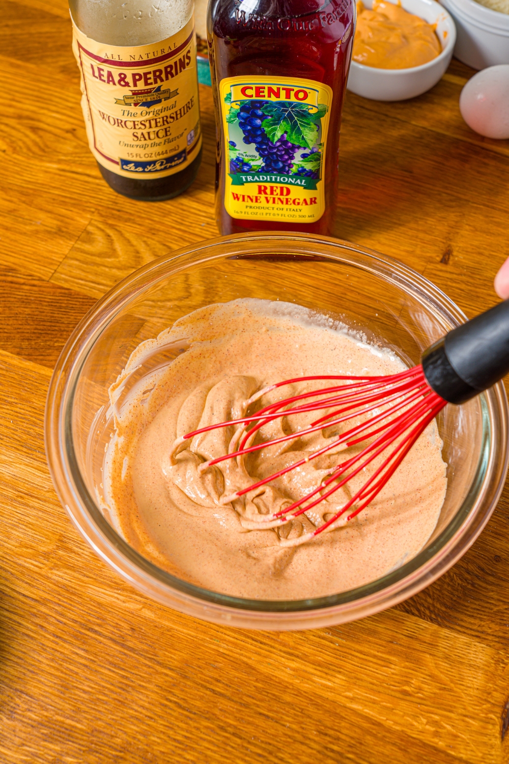 A glass bowl with a whisk mixing ingredients to make crab cake sauce including mayo, dijon mustard, and seasonings. The bowl is on a wooden counter.