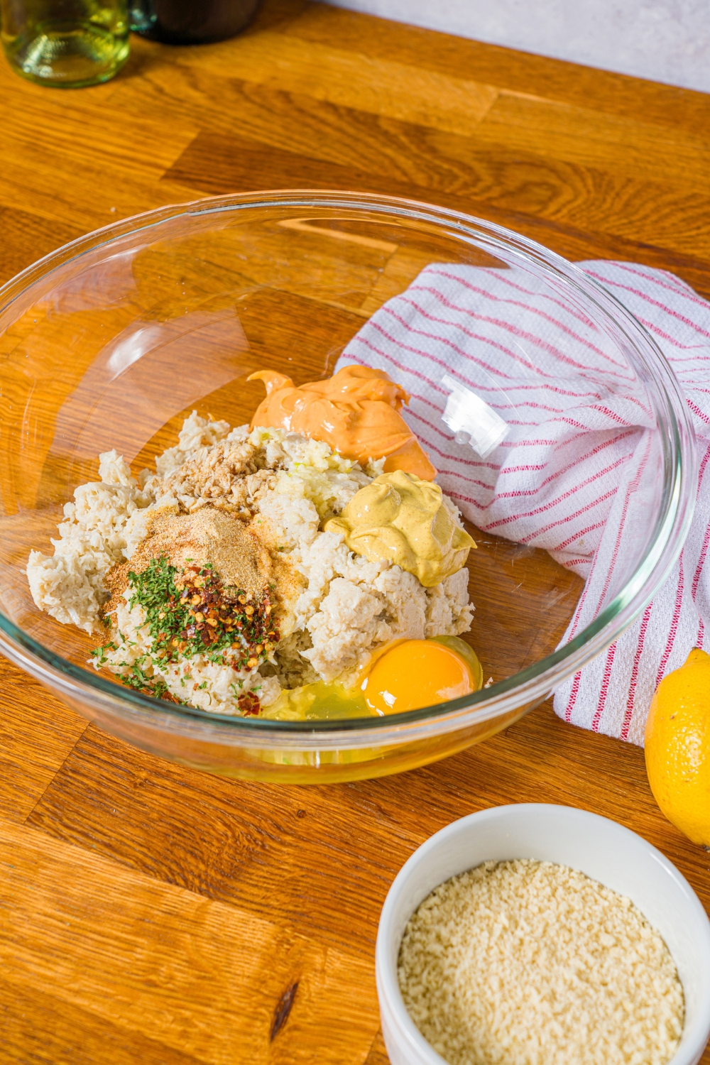 A large bowl with ingredients to make crab cakes including lump crab, mustard, spicy mayo, egg, and seasonings. The bowl is on a wooden counter with a red striped napkin.