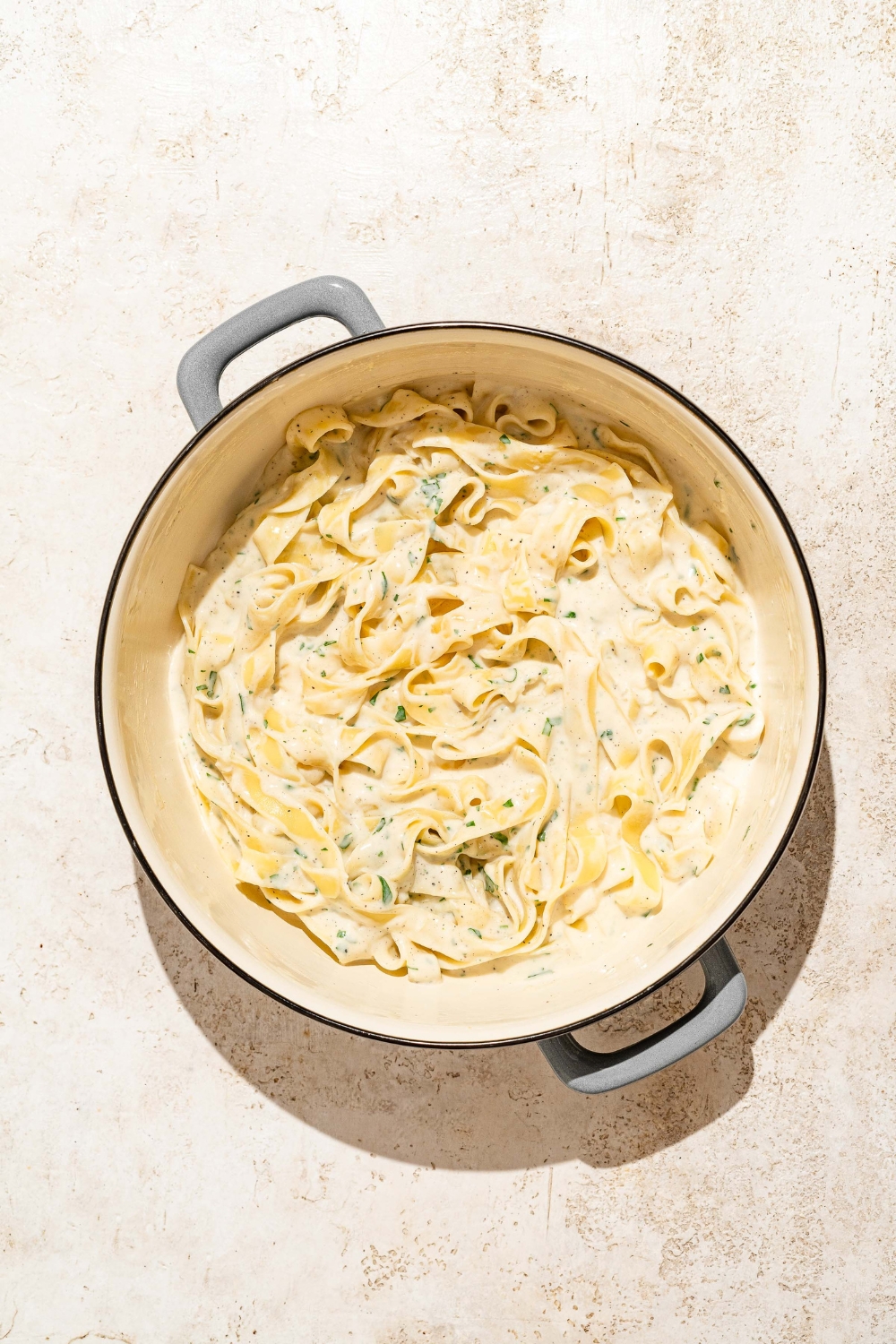 A large pot with fettuccine pasta tossed in alfredo sauce and seasonings. The pot is on a tan counter.