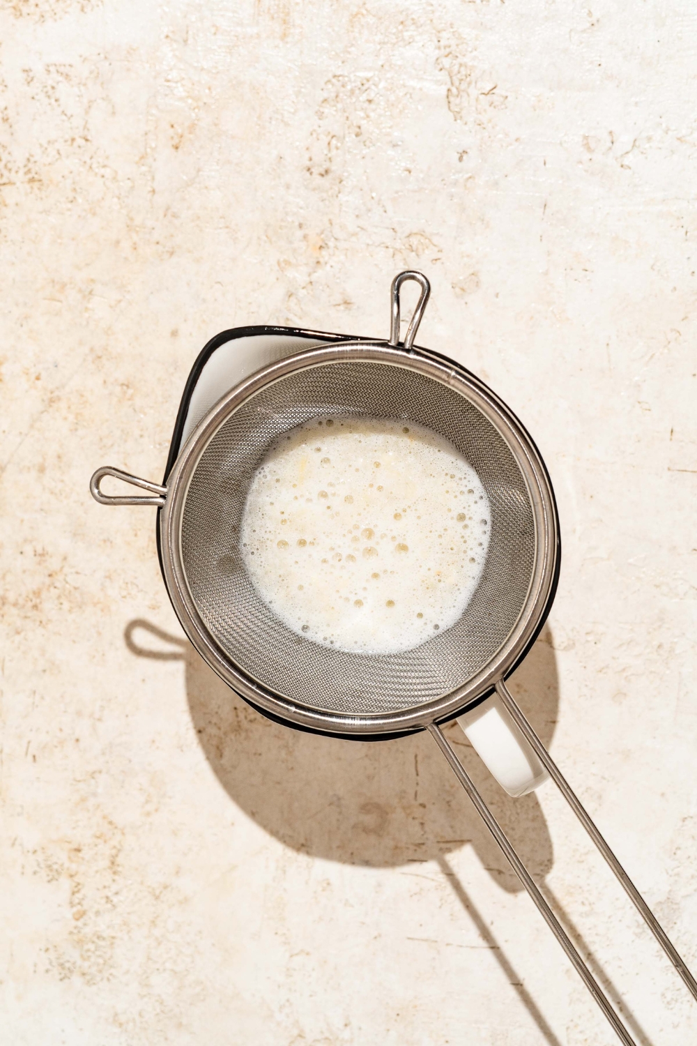 A mesh sieve straining garlic water over a bowl. The bowl is on a tan counter.