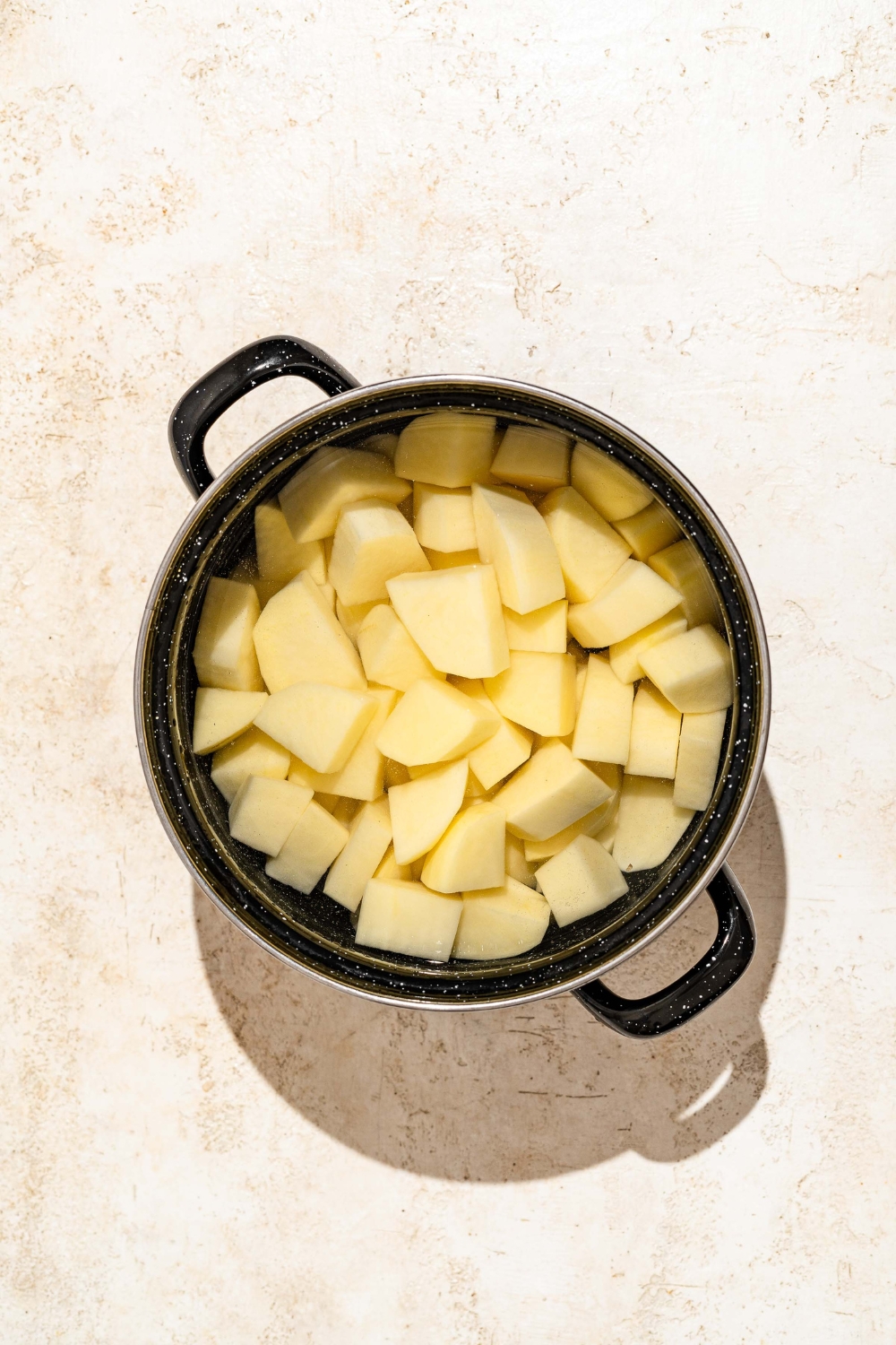 A large stock pot with sliced potatoes in water. The pot is on a tan counter.