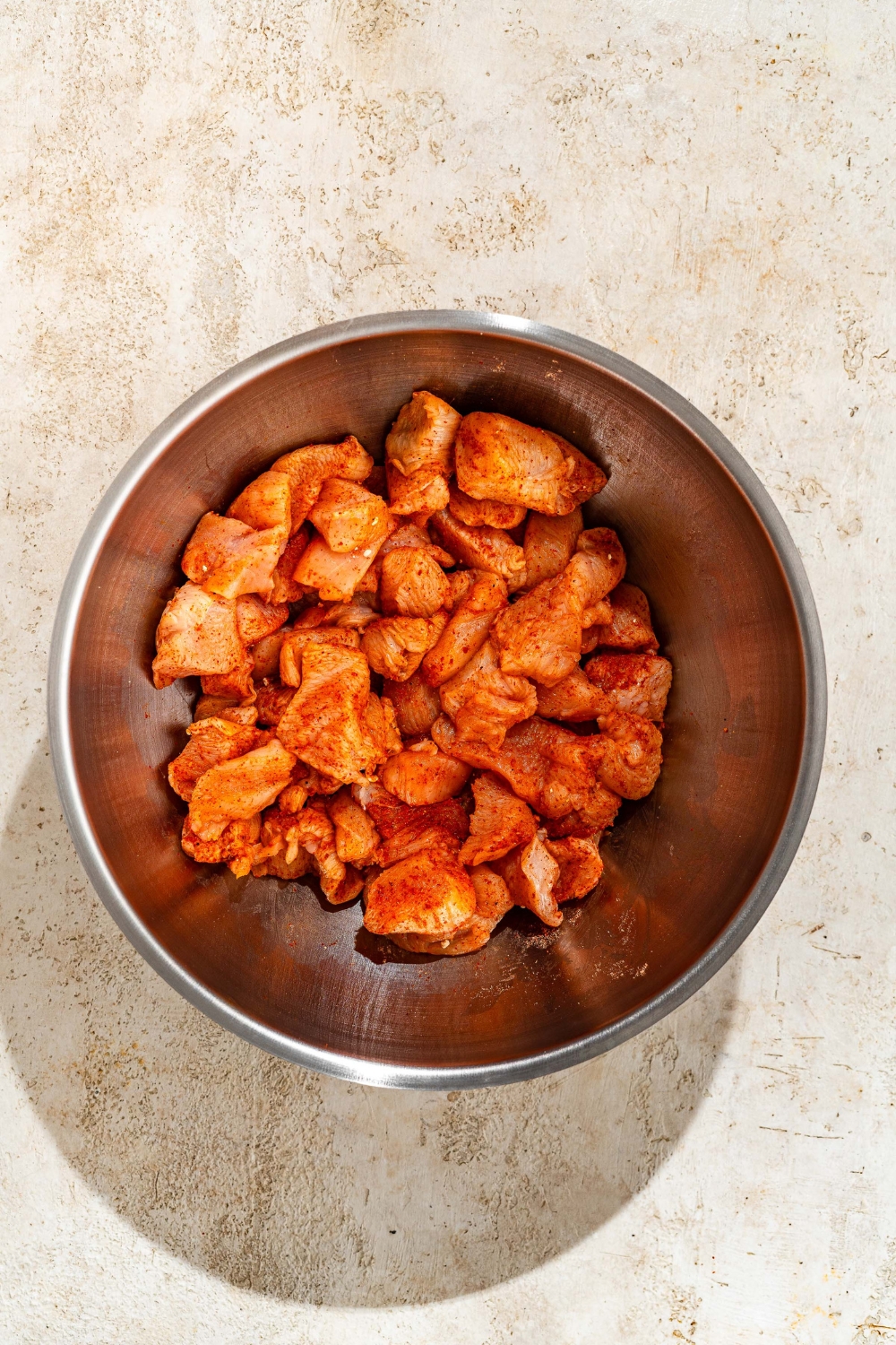 A large mixing bowl with chicken tossed in seasonings. The bowl is on a tan counter.