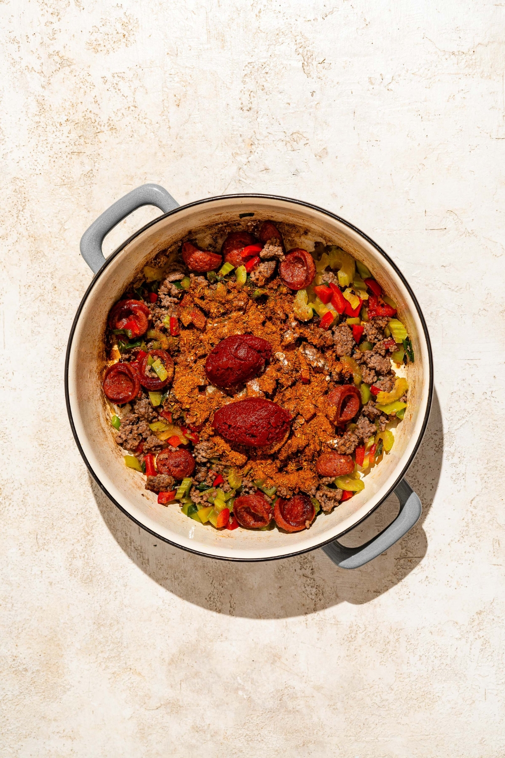 A dutch oven with sautéed sausage, ground beef, and vegetables topped with tomato paste and seasonings. The dutch oven is on a tan counter.