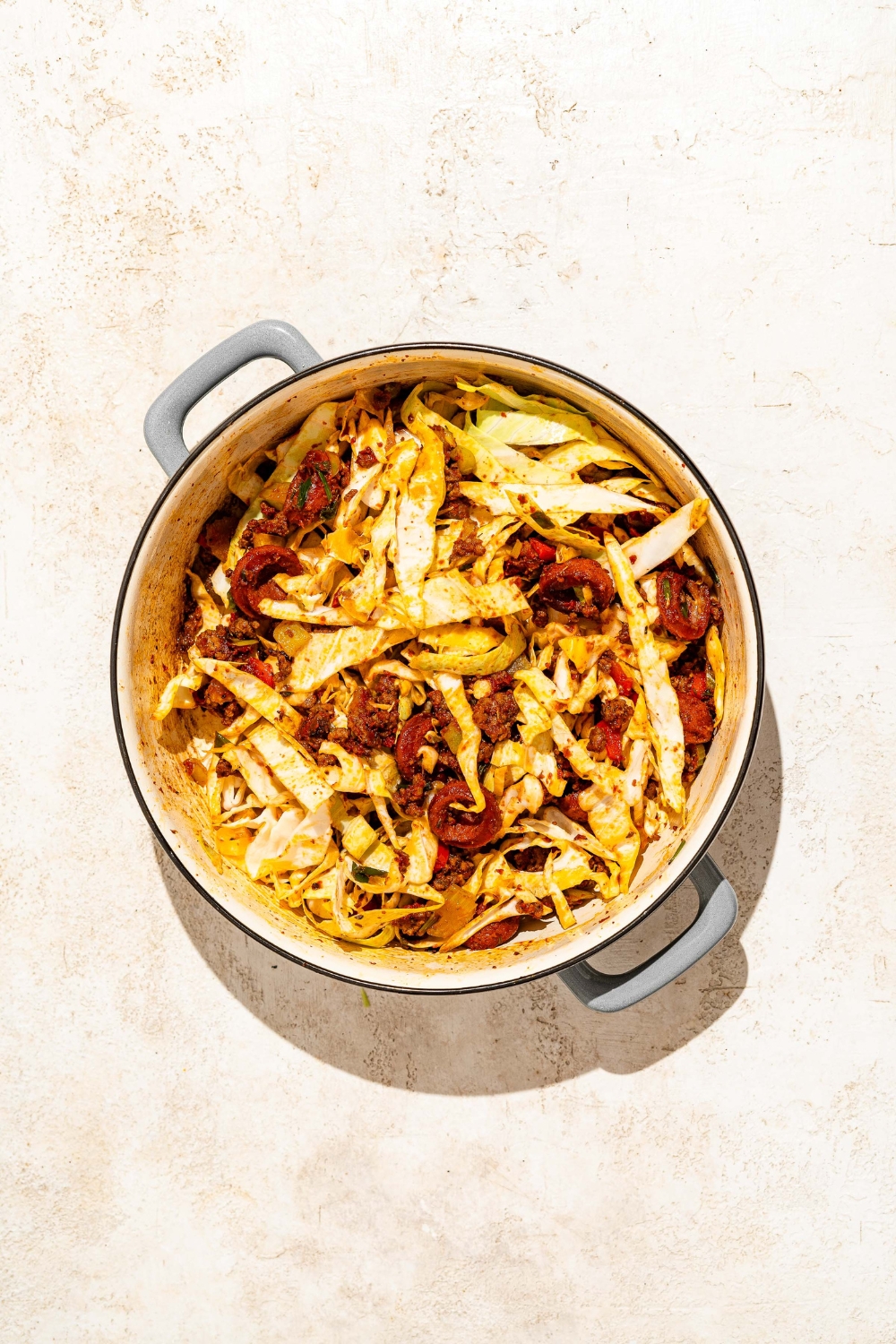 A dutch oven with sautéed sausage, ground beef, and vegetables with sliced cabbage. The dutch oven is on a tan counter.