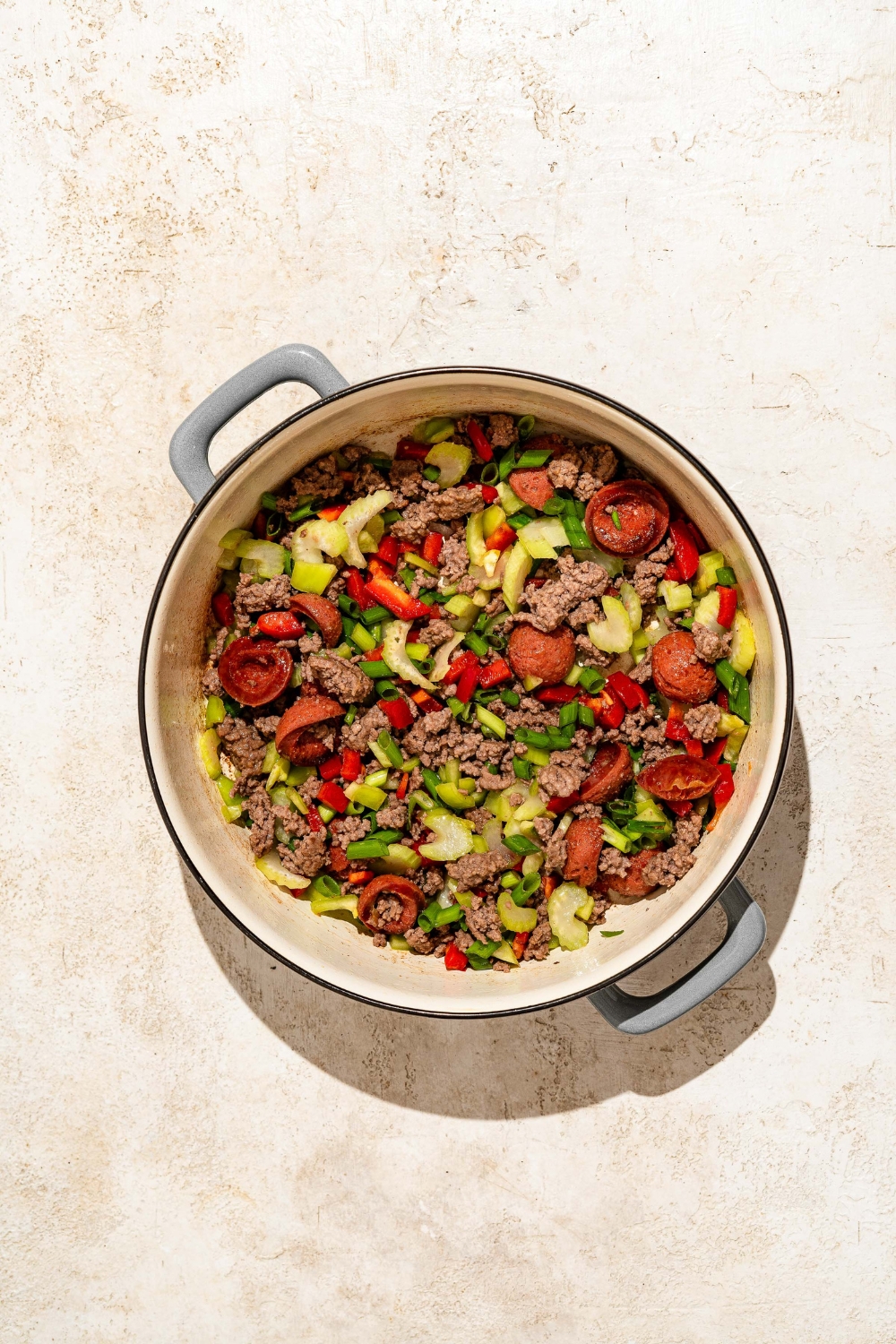 A dutch oven with sautéed ground beef, sausage, and vegetables. The dutch oven is on a tan counter.