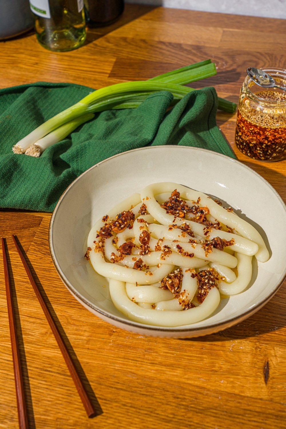 A bowl with spicy potato noodles topped with chili crisp. The bowl is on a wooden counter with a pair of chopsticks, jar of chili crisp, and a green cloth napkin.