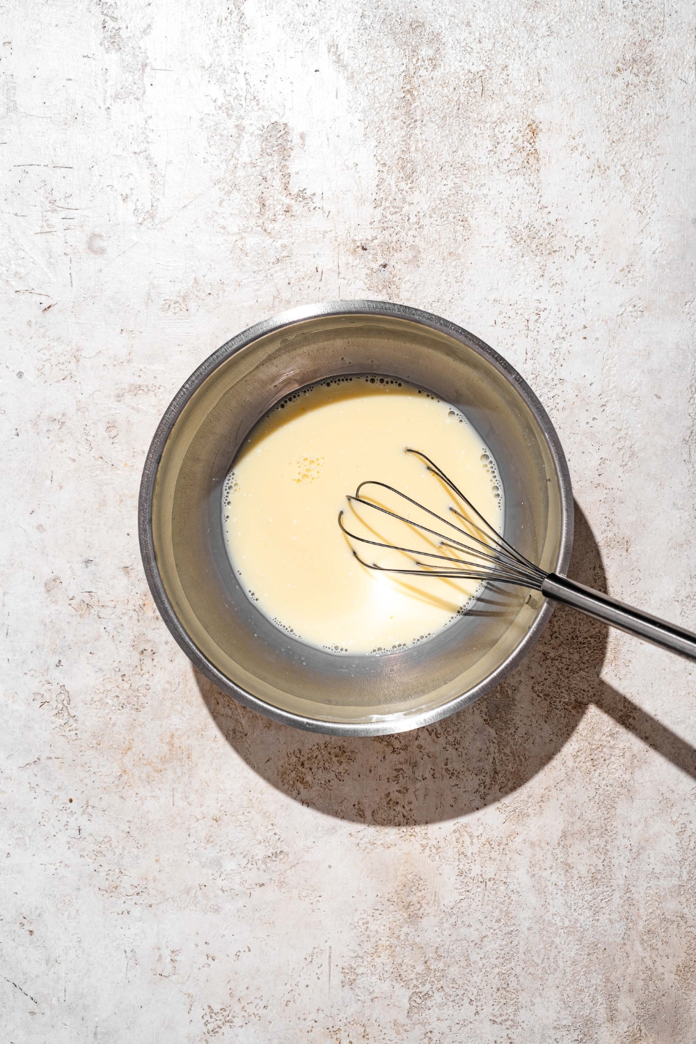 A large bowl with a whisk mixing wet ingredients for funnel cake batter. The bowl is on a tan counter.