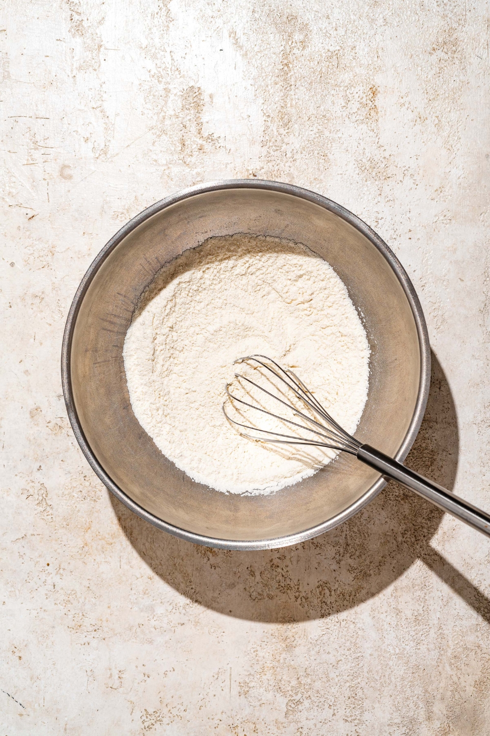 A large bowl with a whisk mixing dry ingredients to make funnel cake batter. The bowl is on a tan counter.