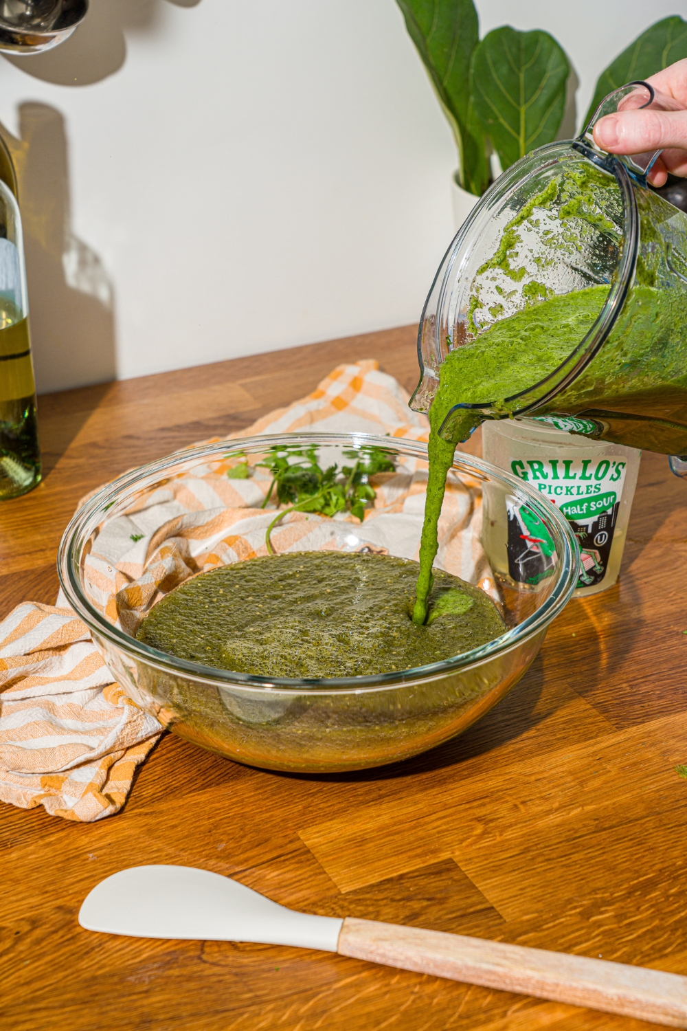 A blender jar adding blended pickles to a glass bowl with a salsa base. The bowl is on a wooden counter with a spatula.