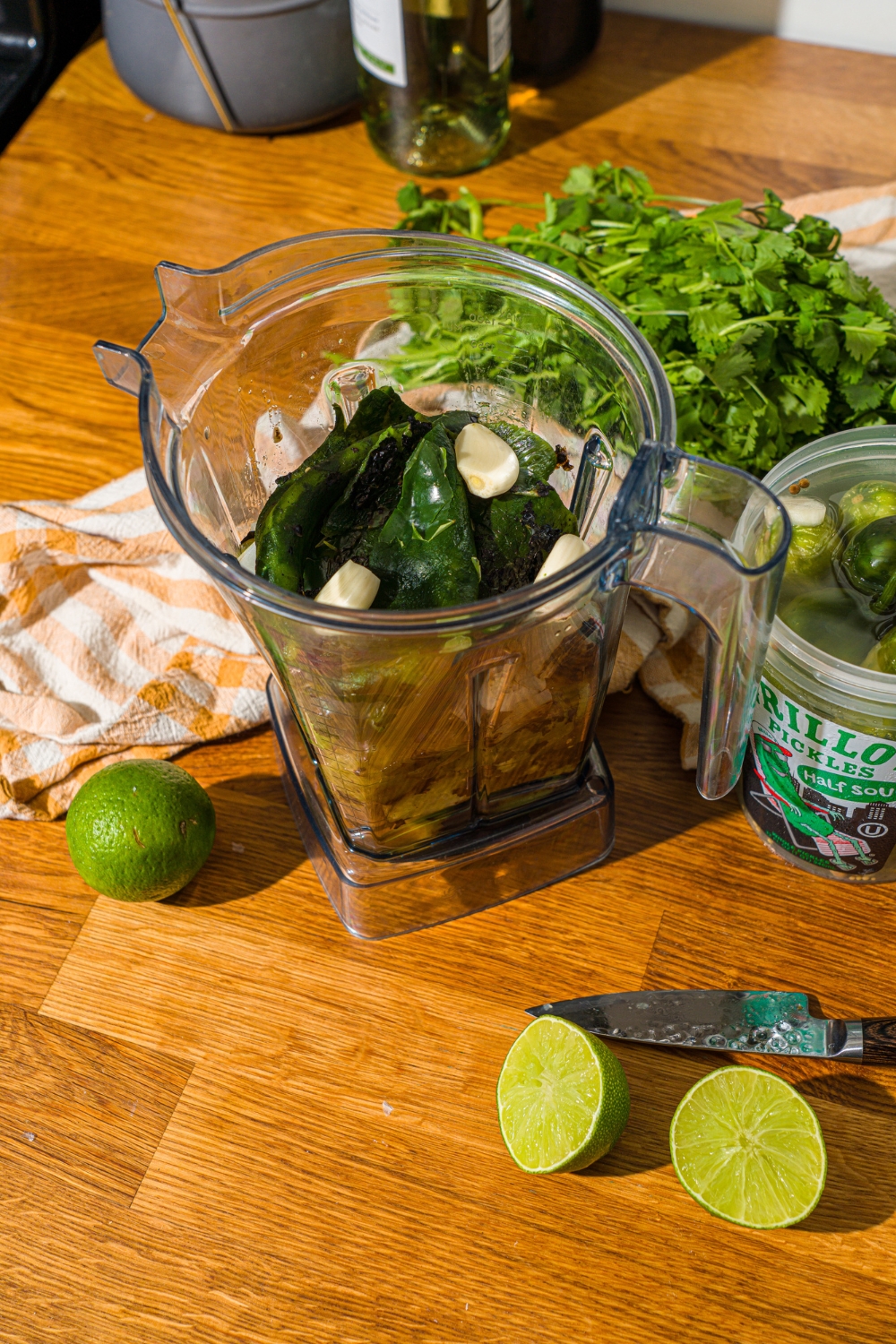 A blender with garlic, poblano peppers, tomatillos, and onion. The blender is on a wooden counter with limes and a container of Grillo's pickles.
