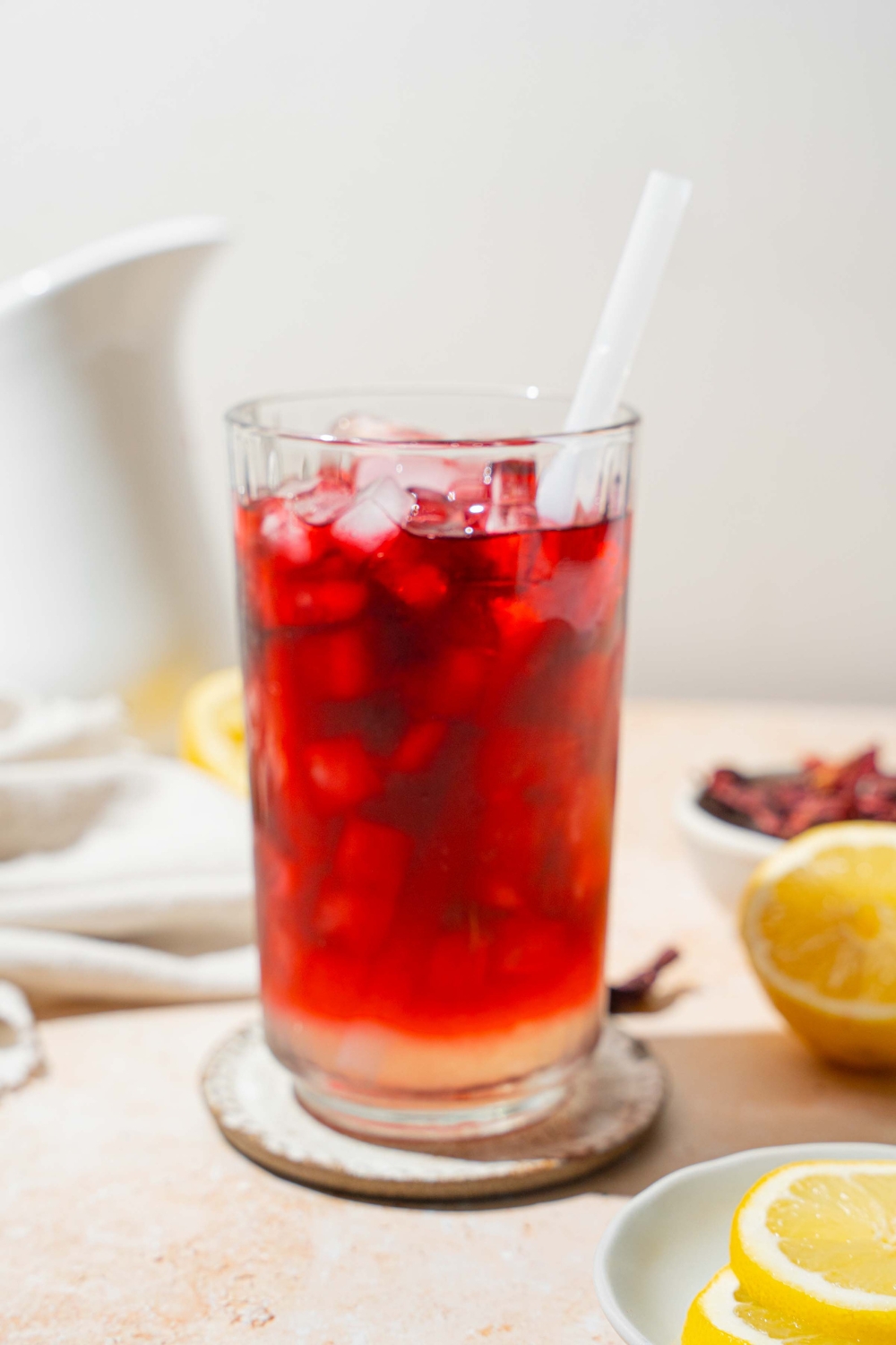 A tall glass of hibiscus lemonade served with ice and a straw. The glass is on a coaster on a tan counter with a white cloth napkin, sliced lemon, and a bowl of hibiscus tea leaves.