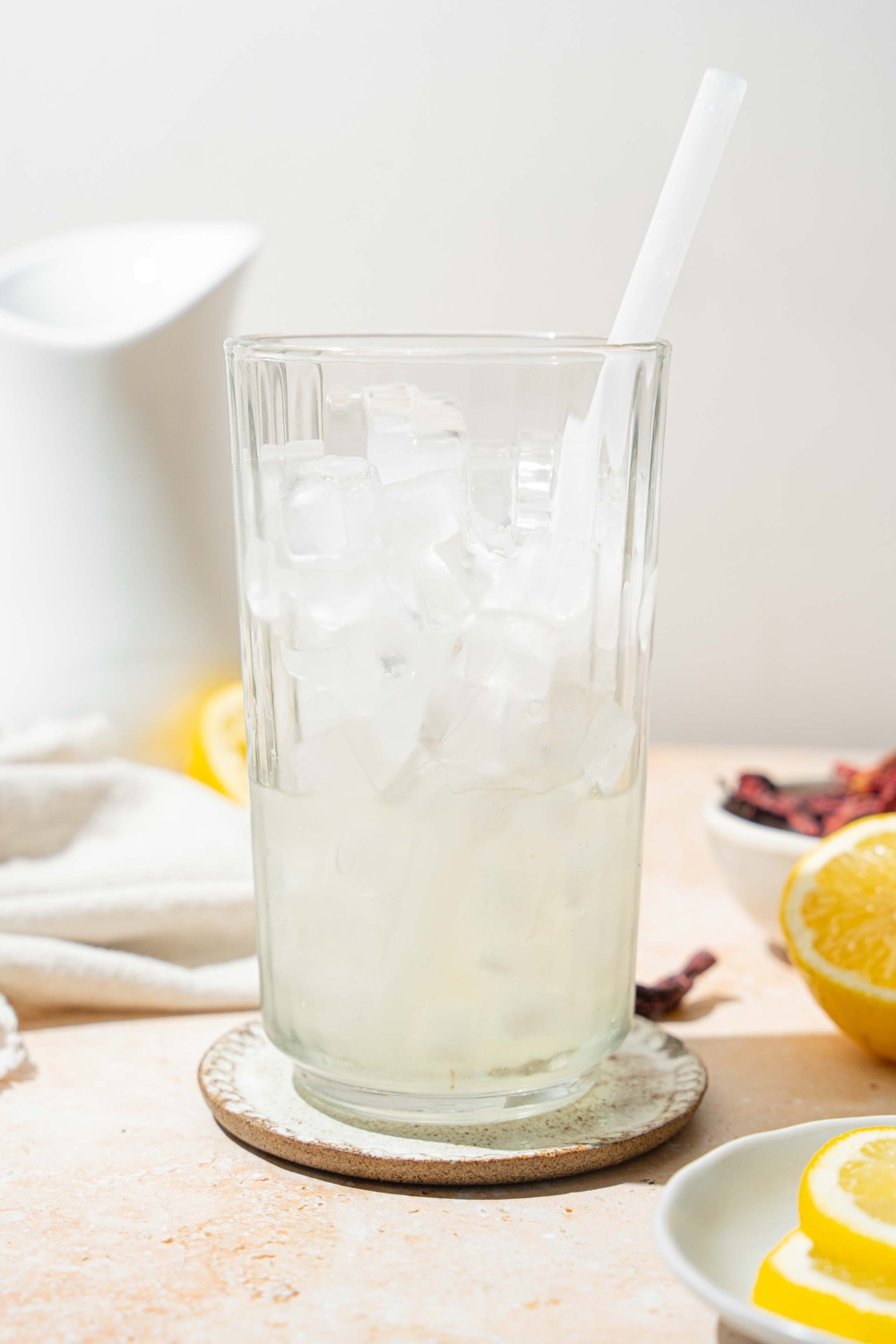 A tall glass with ice, lemonade, and a straw. The glass is on a coaster on a tan counter with a white cloth napkin, sliced lemon, and a bowl of hibiscus tea leaves.