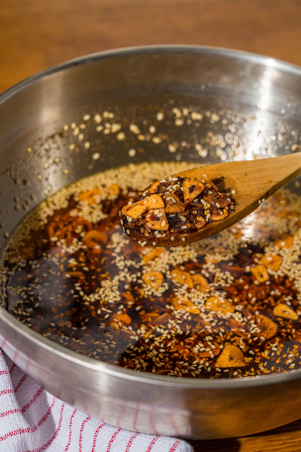 A wooden spoon dipped in a large bowl of chili crisp oil. The bowl is on a wooden counter with a white striped napkin.