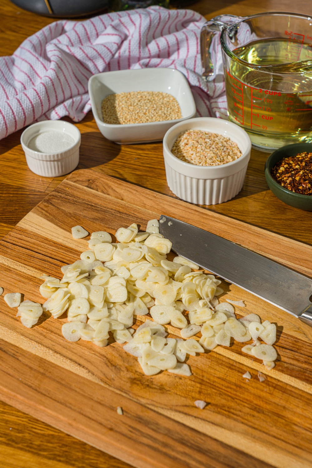 A wooden board with sliced garlic and a knife. The board is on a wooden counter with bowls of ingredients.