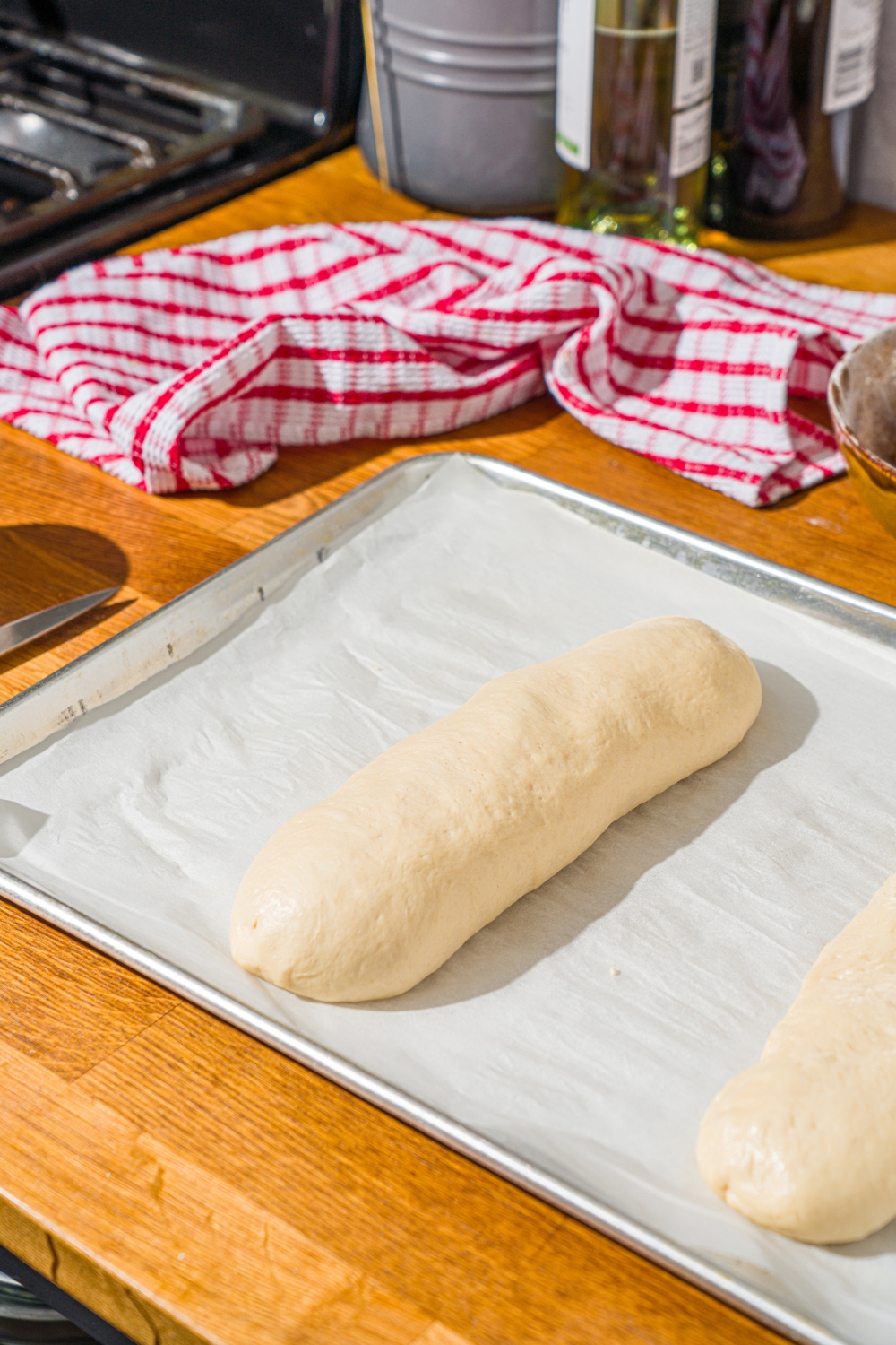 A baking sheet lined with parchment paper with unbaked homemade hoagie rolls. The baking sheet is on a wooden counter with a red striped napkin.