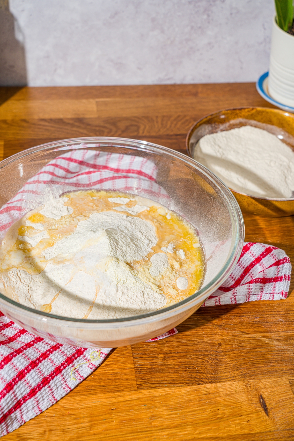 A large glass bowl with flour, melted butter, and milk powder added to a yeast mixture. The bowl is on a wooden counter with a red striped napkin.
