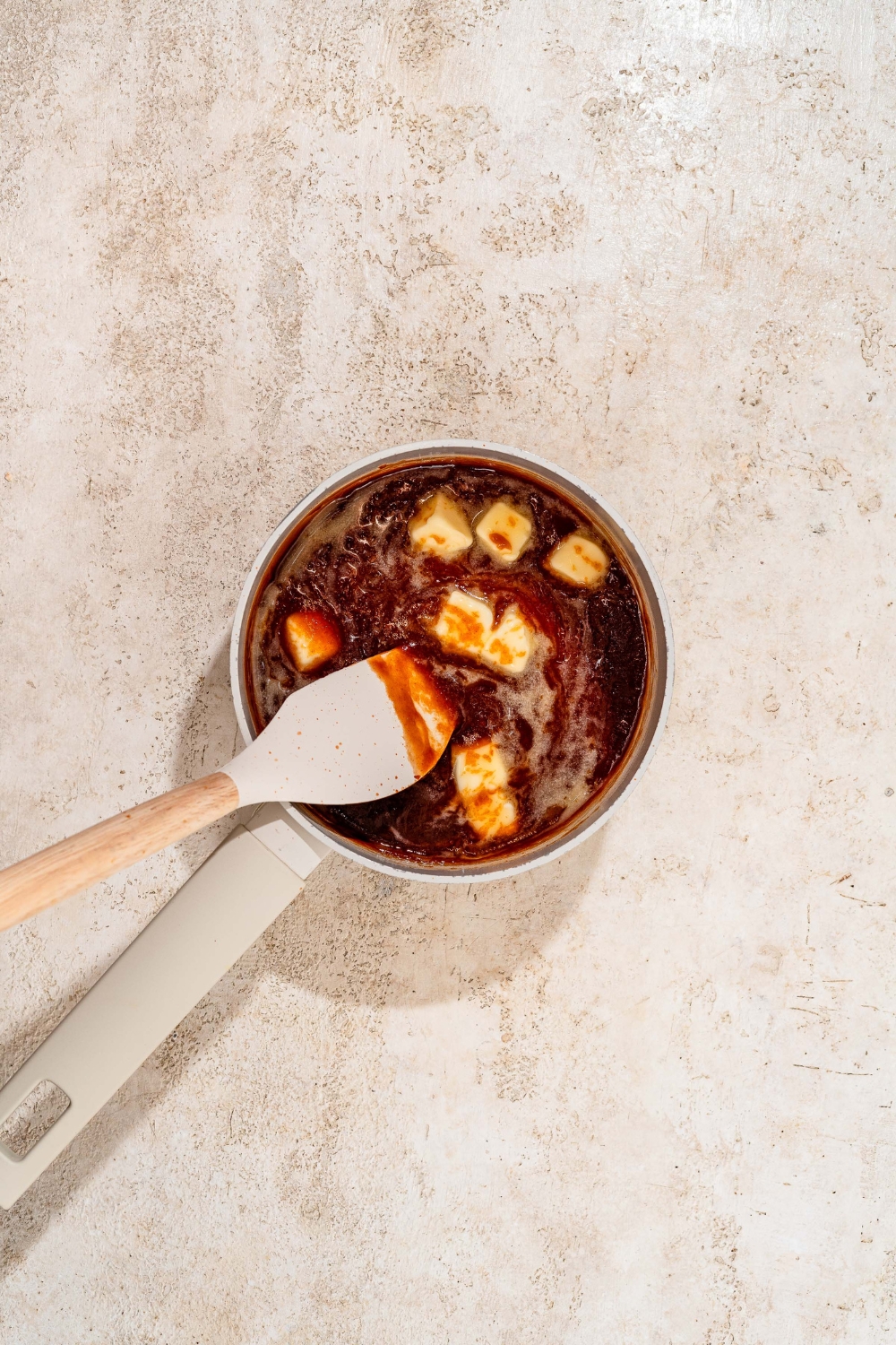 A sauce pan with a spatula mixing a honey BBQ sauce with butter. The pot is on a tan counter.