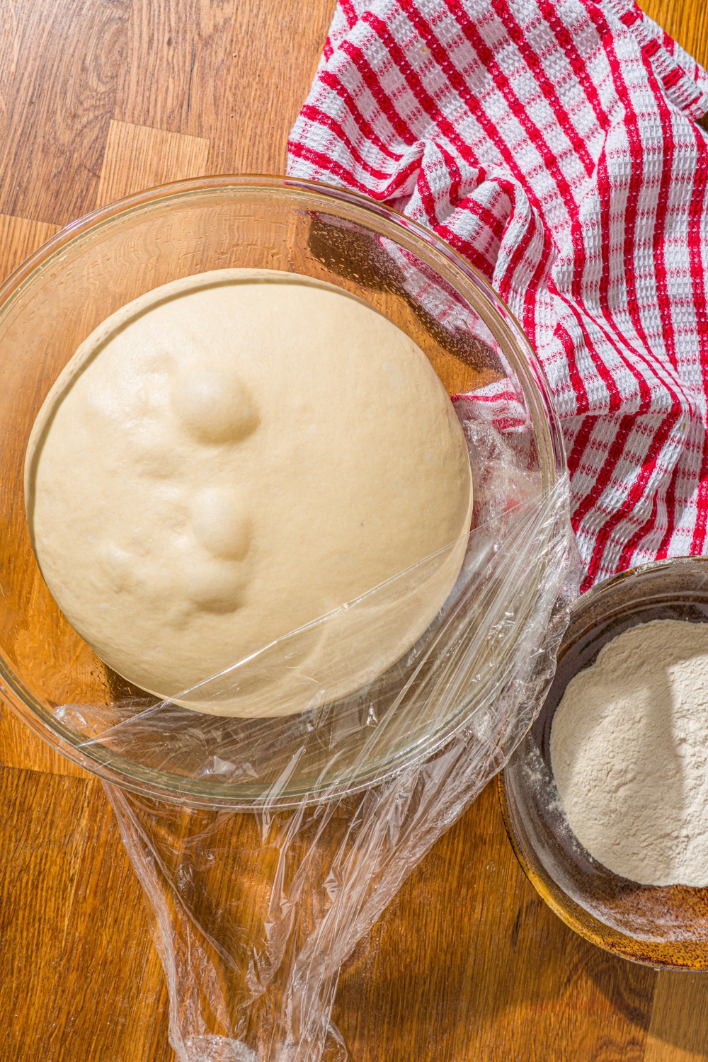 A glass bowl with a ball of bread dough with part of the bowl wrapped in saran wrap. The bowl is on a wooden counter with a red striped napkin.