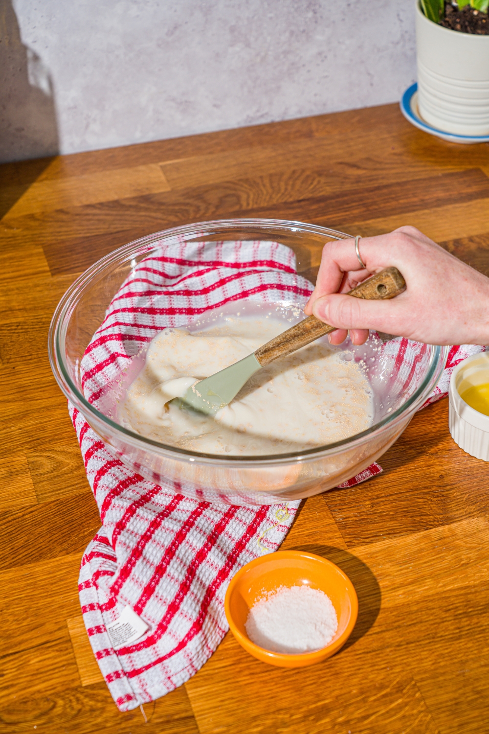 A glass bowl with a spatula mixing a yeast mixture with water, milk, and honey. The bowl is on a wooden counter with a red striped napkin.