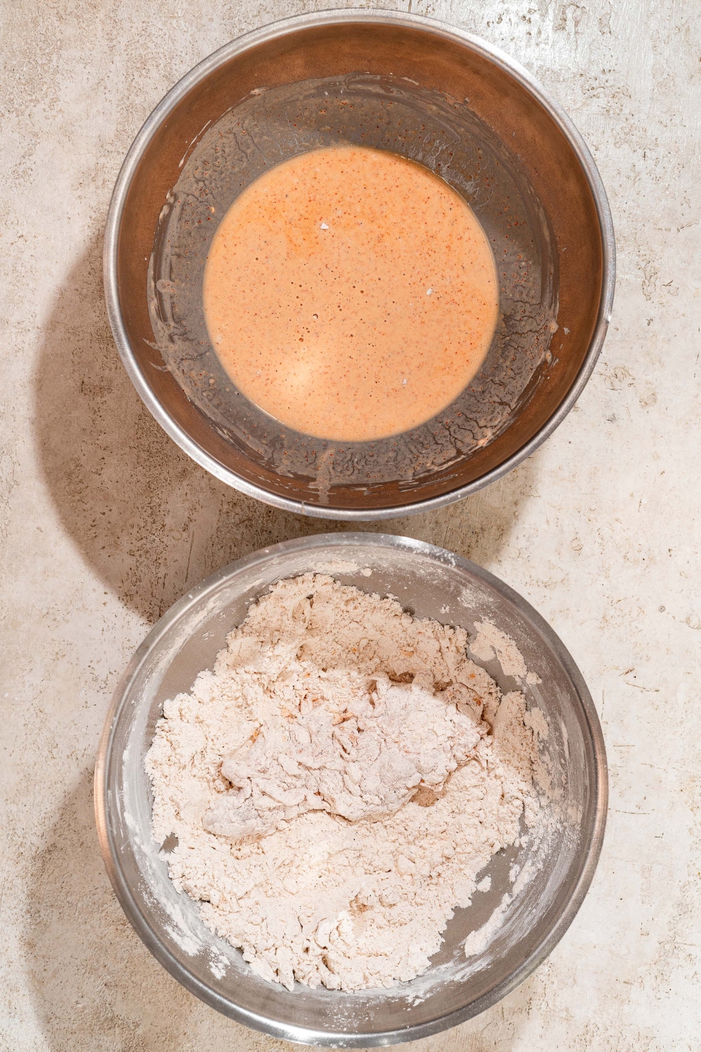 One large bowl of wet batter and another large bowl of seasoned flour with a piece of marinated chicken tender. The bowls are on a tan counter.