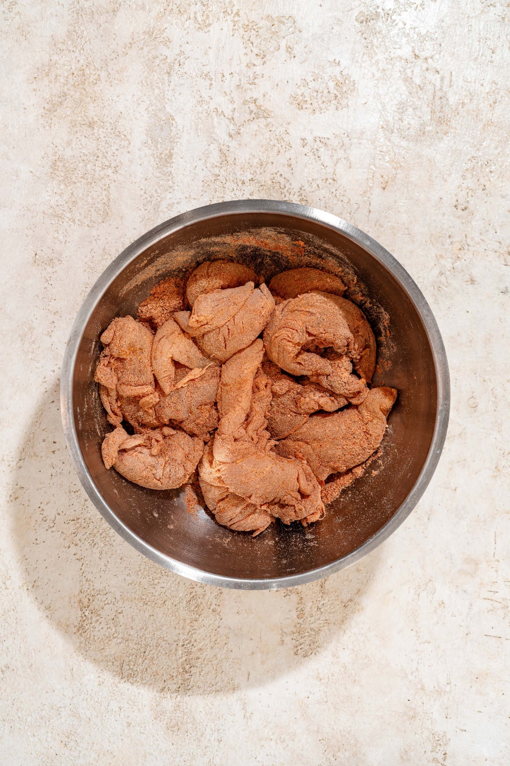 A large bowl with uncooked chicken tenders marinating in a dry mixture. The bowl is on a tan counter.