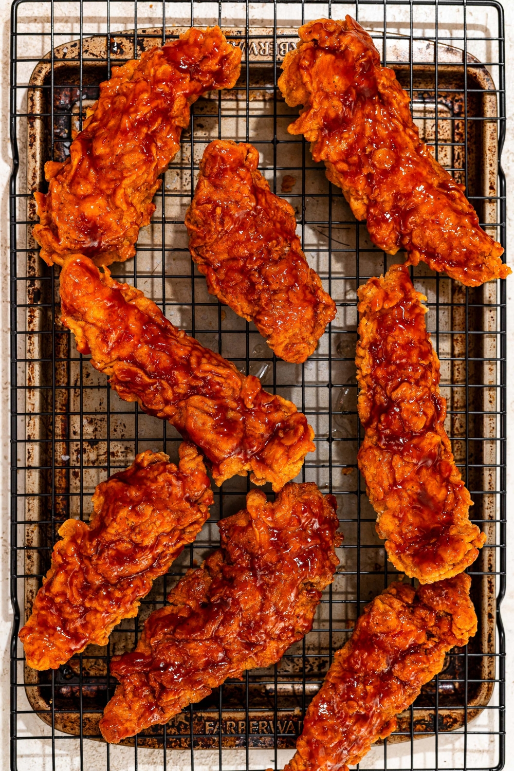 A wire rack with fried chicken tenders coated in honey BBQ sauce. The rack is on a baking sheet on a tan counter.