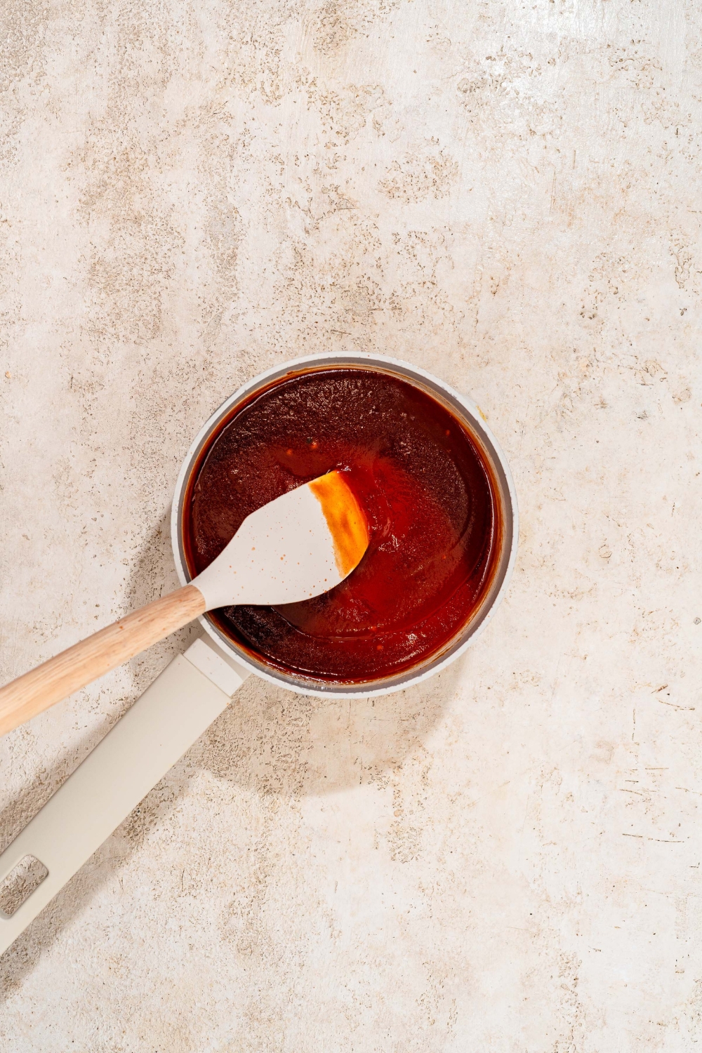 A saucepan with a spatula mixing ingredients to make honey BBQ sauce. The pot is on a tan counter.
