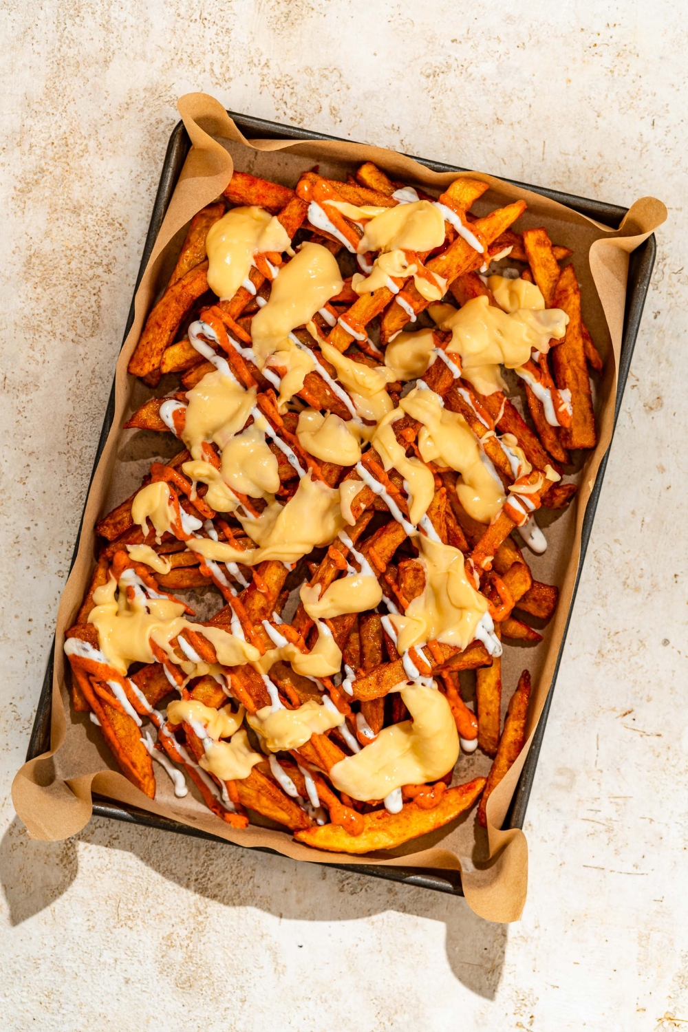 A baking sheet lined with parchment paper with fries toped with cheese sauce and yogurt sauce. The sheet is on a tan counter.
