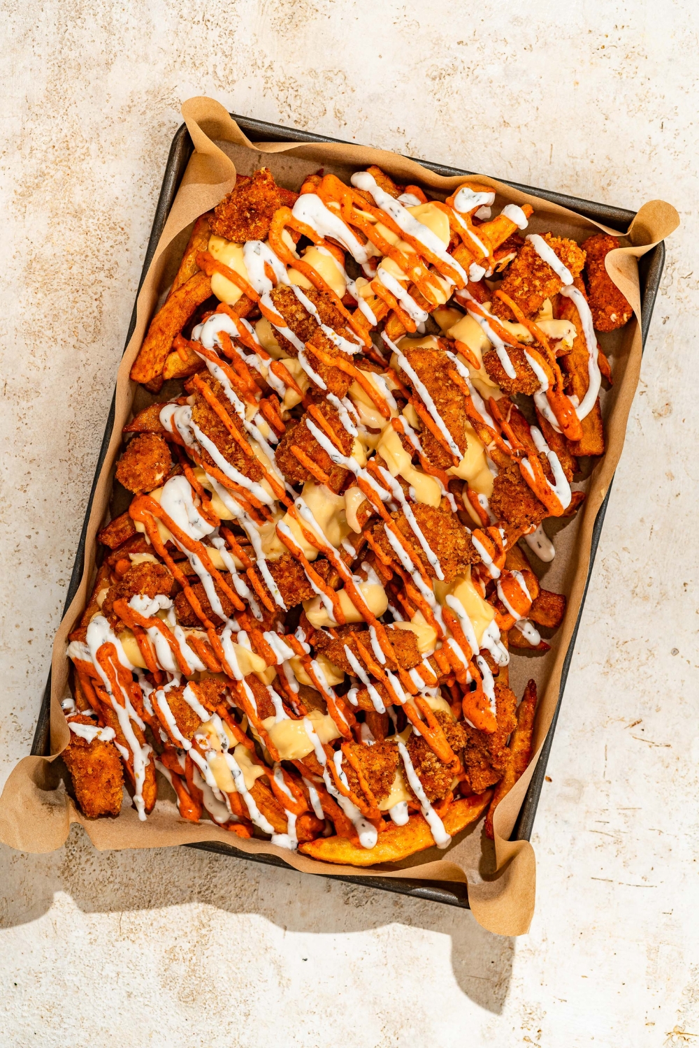 A baking sheet lined with parchment paper with loaded fries topped with crispy chicken, sriracha mayo sauce, yogurt sauce, and cheese sauce. The sheet is on a tan counter.
