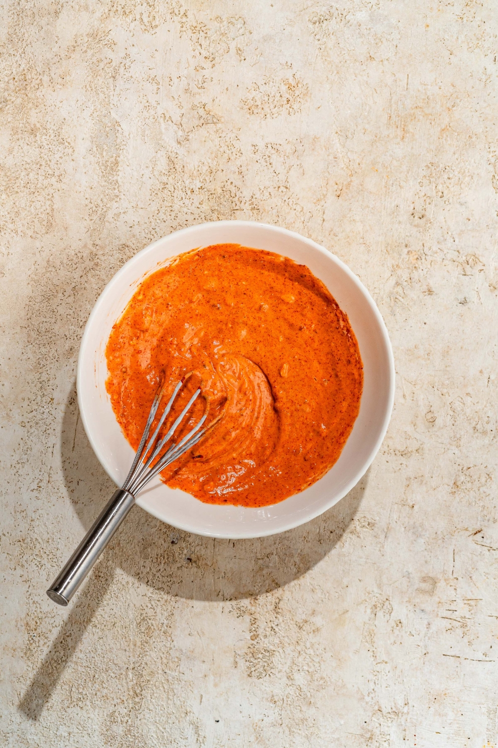 A white bowl with a whisk mixing ingredients to make a sriracha mayo sauce. The bowl is on a tan counter.