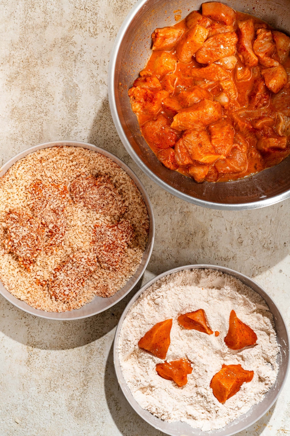 A tan counter with three large bowls for breading seasoned chicken. One bowl contains seasoned chicken mixed with egg, the second bowl contains flour, and the third bowl contains panko breadcrumbs.