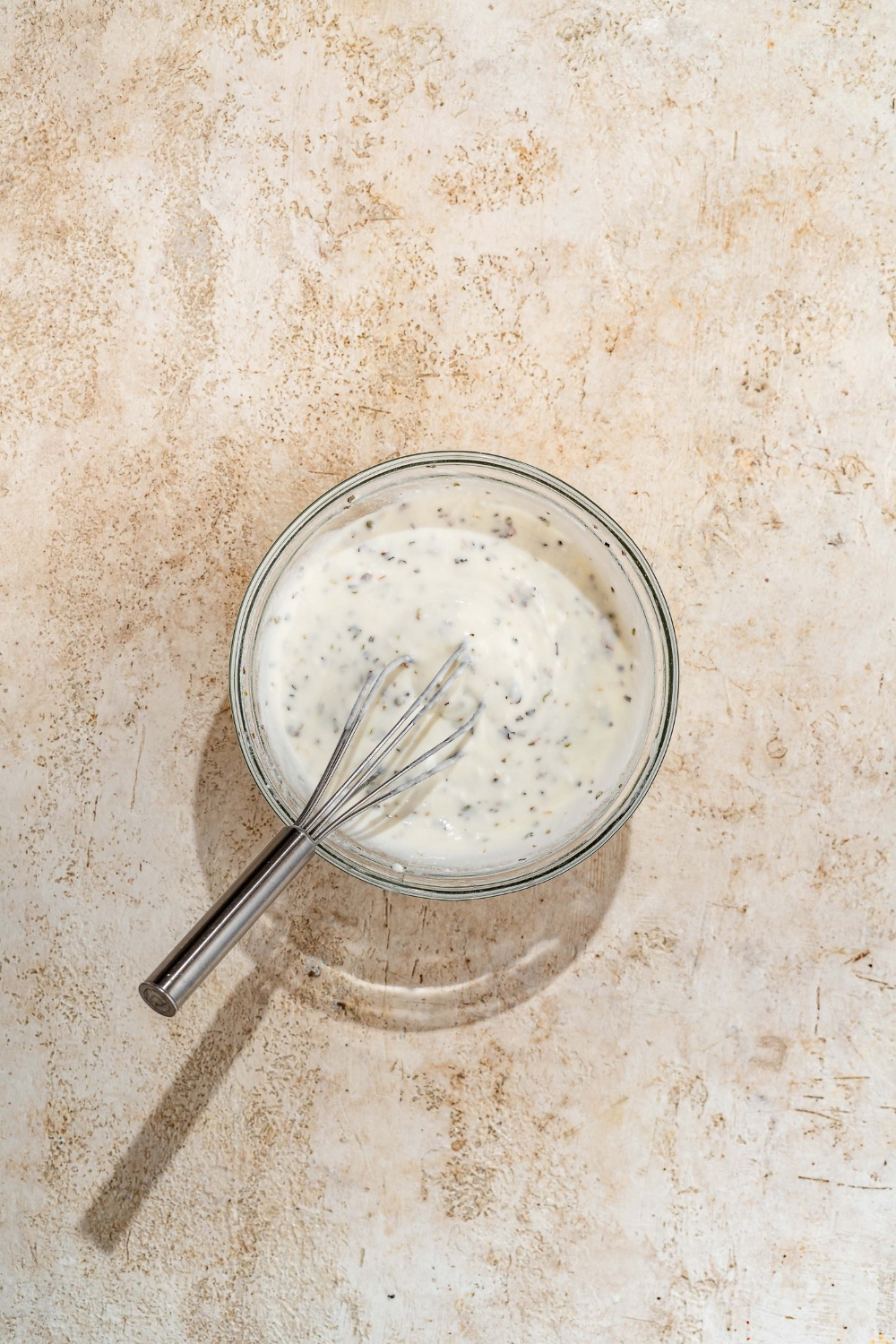 A small glass bowl with a whisk mixing ingredients to make yogurt sauce including yogurt, mayo, and herbs.