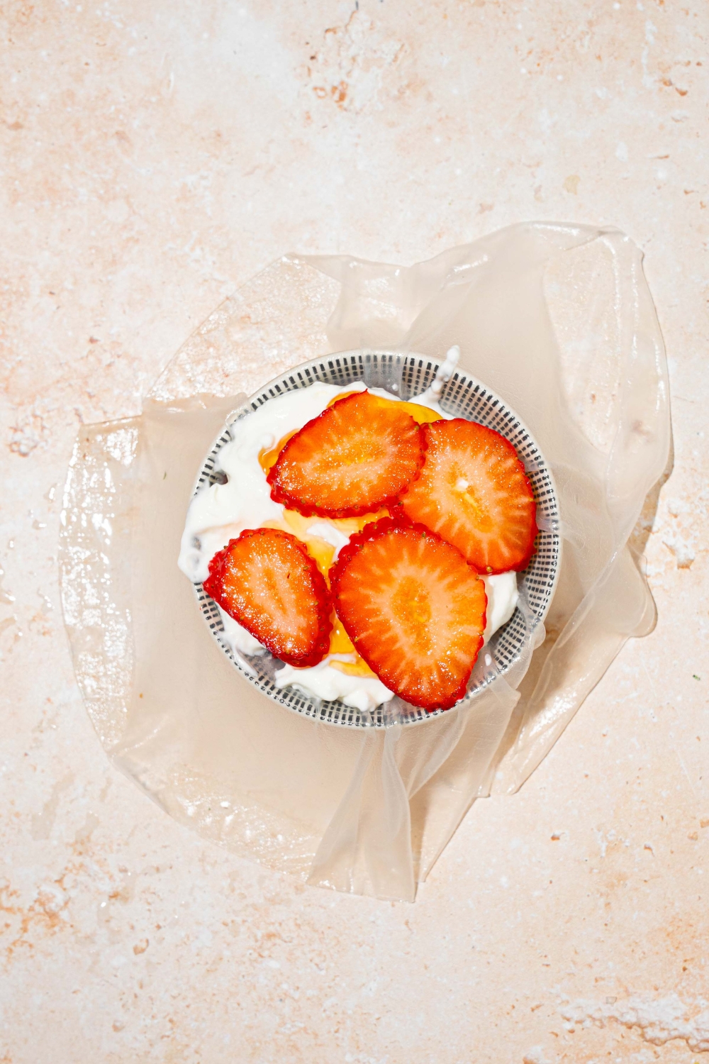 A cup with rice paper filled with yogurt, honey, and sliced strawberries. The cup is on a tan counter.