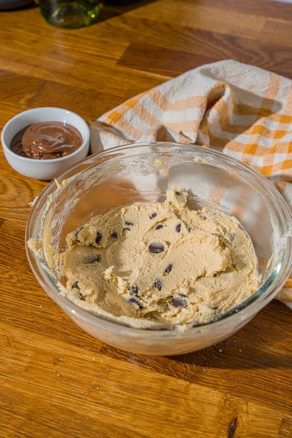 A glass bowl with cookie dough mixed with chocolate chips. The bowl is on a wooden counter with a bowl of nutella and yellow striped napkin.