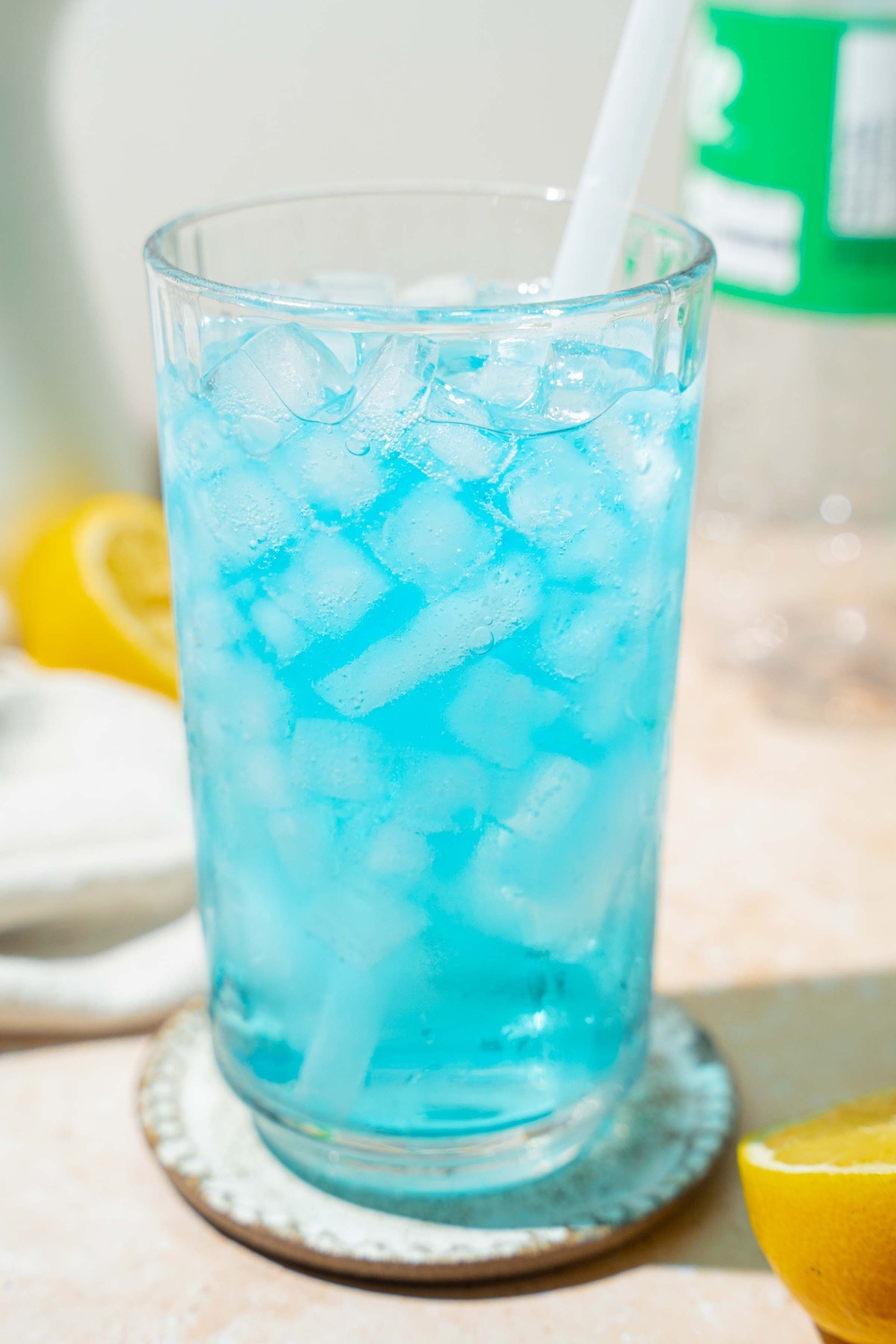 A tall glass with ocean water drink served with pebble ice and a straw. The glass is on a coaster on a an counter with sliced lemon.