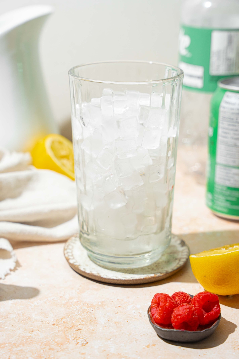 A tall glass filled with pebble ice. The glass is on a coaster on a tan counter with a small bowl of raspberries and sliced lemon.