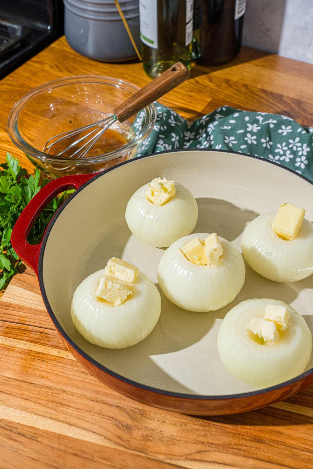 A dutch oven with several onions with the tops sliced off and stuffed with butter. The dutch oven is on a wooden counter with a bowl of seasoned butter and a green floral napkin.