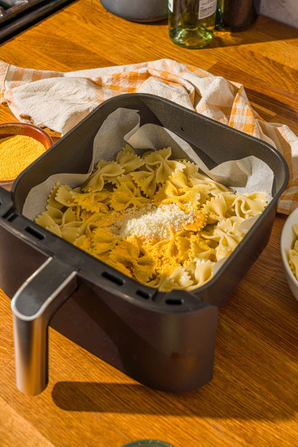 An air fryer lined with parchment paper with cooked bowtie pasta coated in oil and topped with parmesan cheese and mac and cheese powder. The air fryer is on a wooden counter with a yellow striped napkin.