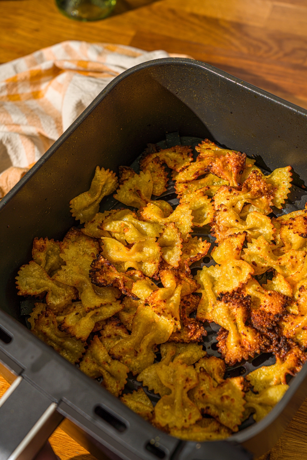 An air fryer basket with air fried bowtie pasta chips seasoned in cheese. The basket is on a wooden counter with a yellow striped napkin.