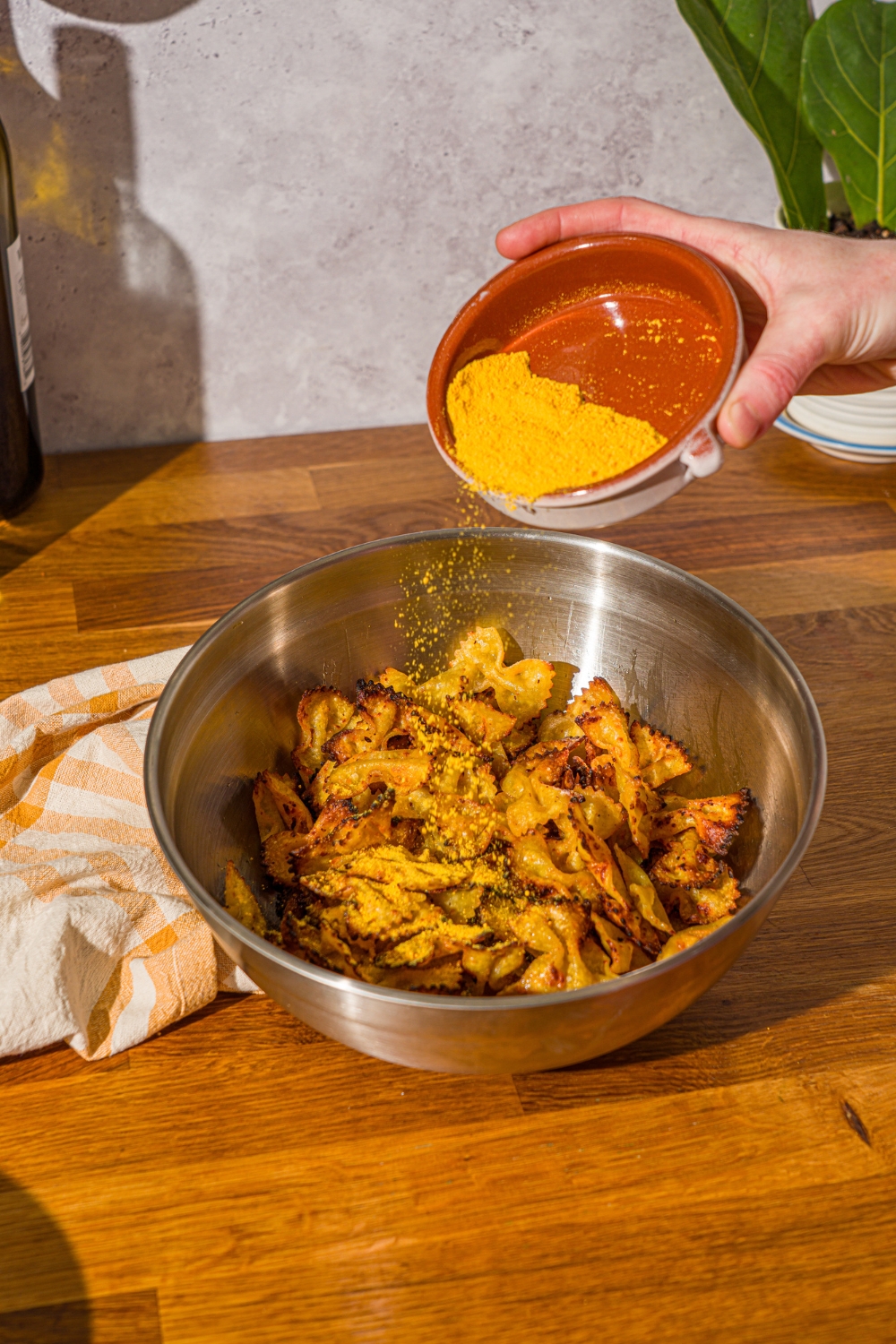 A mixing bowl with air fried bowtie pasta chips with a bowl of mac and cheese powder being poured over the chips. The bowl is on a wooden counter with a yellow striped napkin.