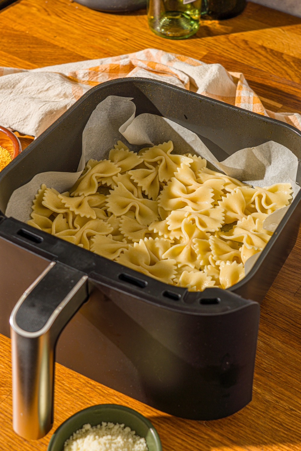 An air fryer basket lined with parchment paper with cooked bowtie pasta. The basket is on a wooden counter with a yellow striped napkin.