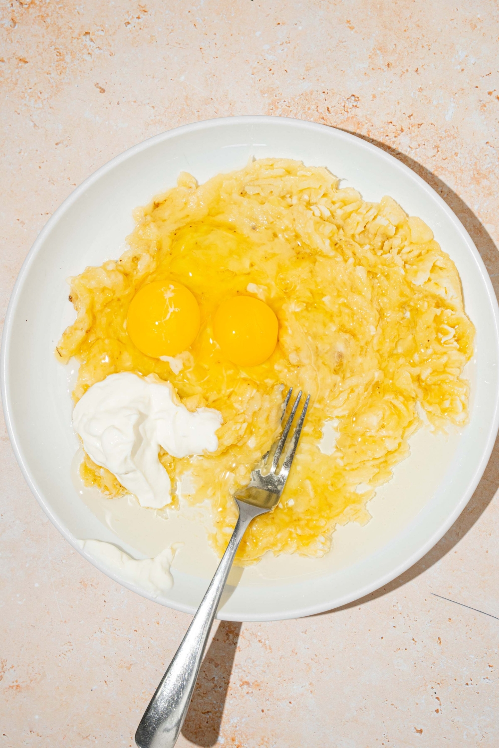 Two eggs and sour cream on top of mashed banana on a plate with a fork.