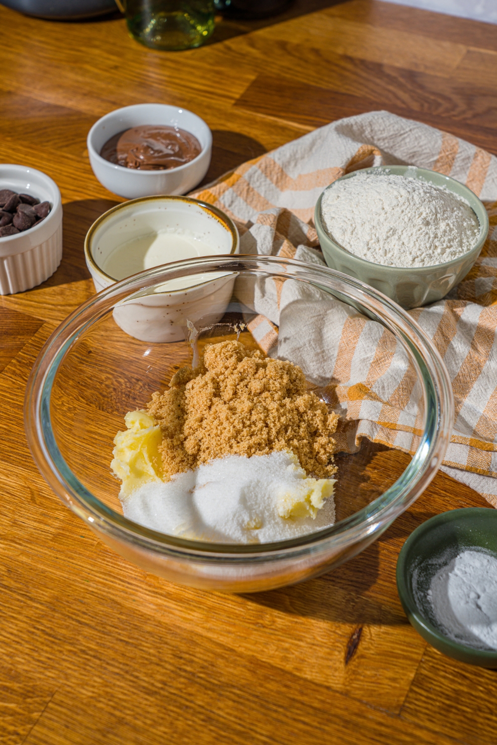 A glass bowl with butter, sugar, and brown sugar. The bowl is on a wooden counter with additional bowls of ingredients.