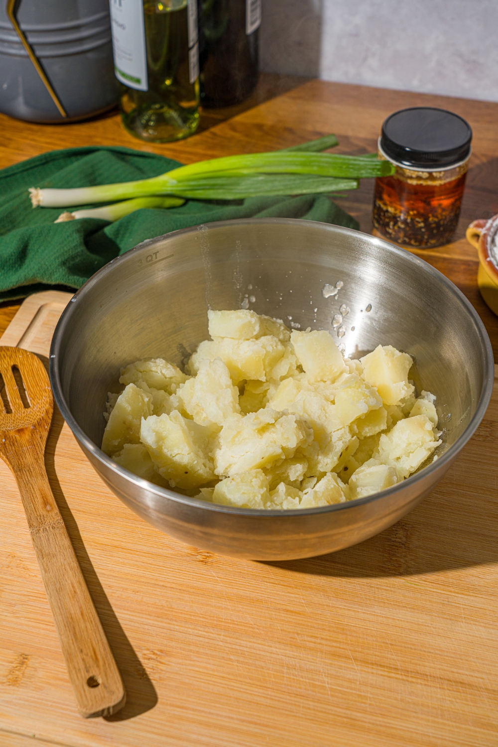 A large mixing bowl with cubed steamed potatoes slightly mashed. The bowl is on a wooden cutting board. The board is on a wooden counter with a green cloth napkin and a wooden spoon.